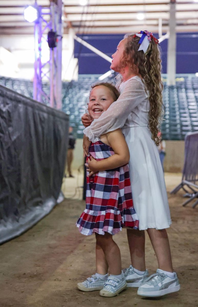 EMBRACING THE YOUTH. A young audience member smiles as her older sister, Haley Jo,  holds her from behind in front of the Montgomery Fairground stadium stage at the Charlie Kirk Memorial Service Sept. 30. Event organizer Charlie Diggs designated Jo to sing the national anthem on stage that night. “Since Kirk always spoke to our young generations,” Diggs said. “I only thought it was right to have them here on stage tonight.”
